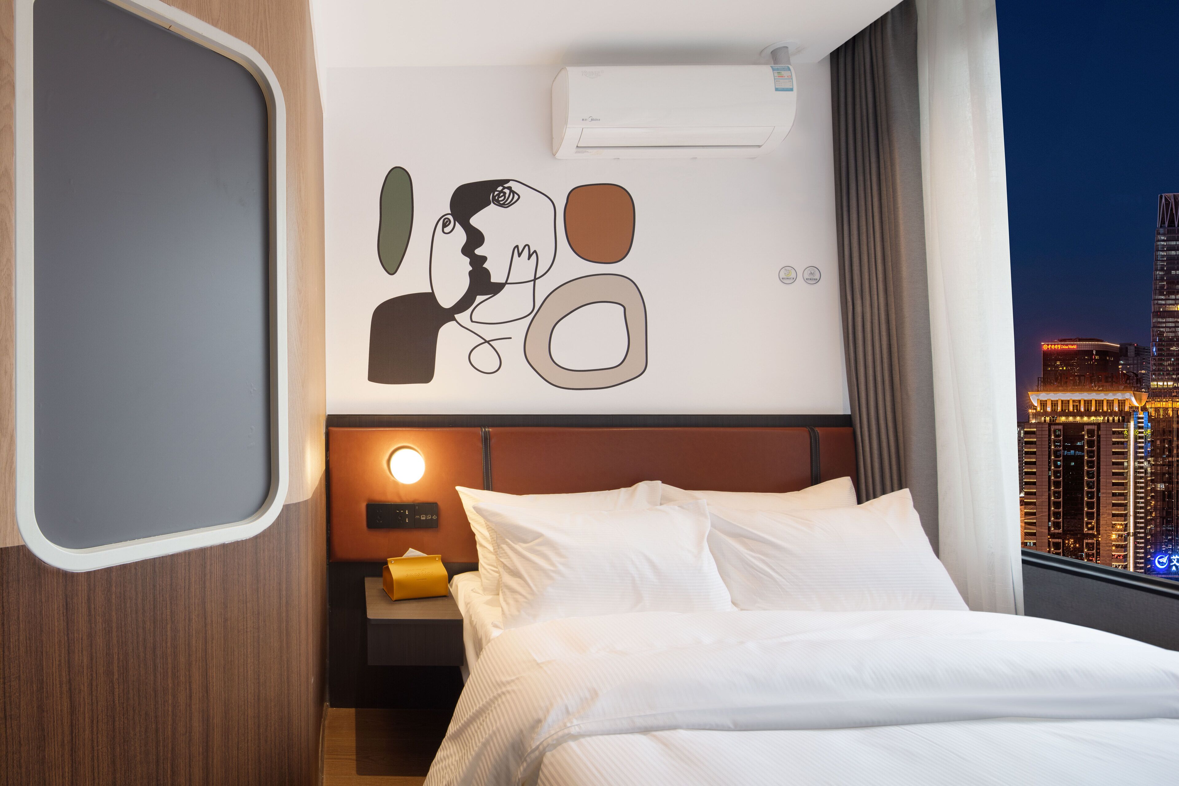 A hotel room with a bed, a nightstand, a wall-mounted air conditioner, and a cityscape view.