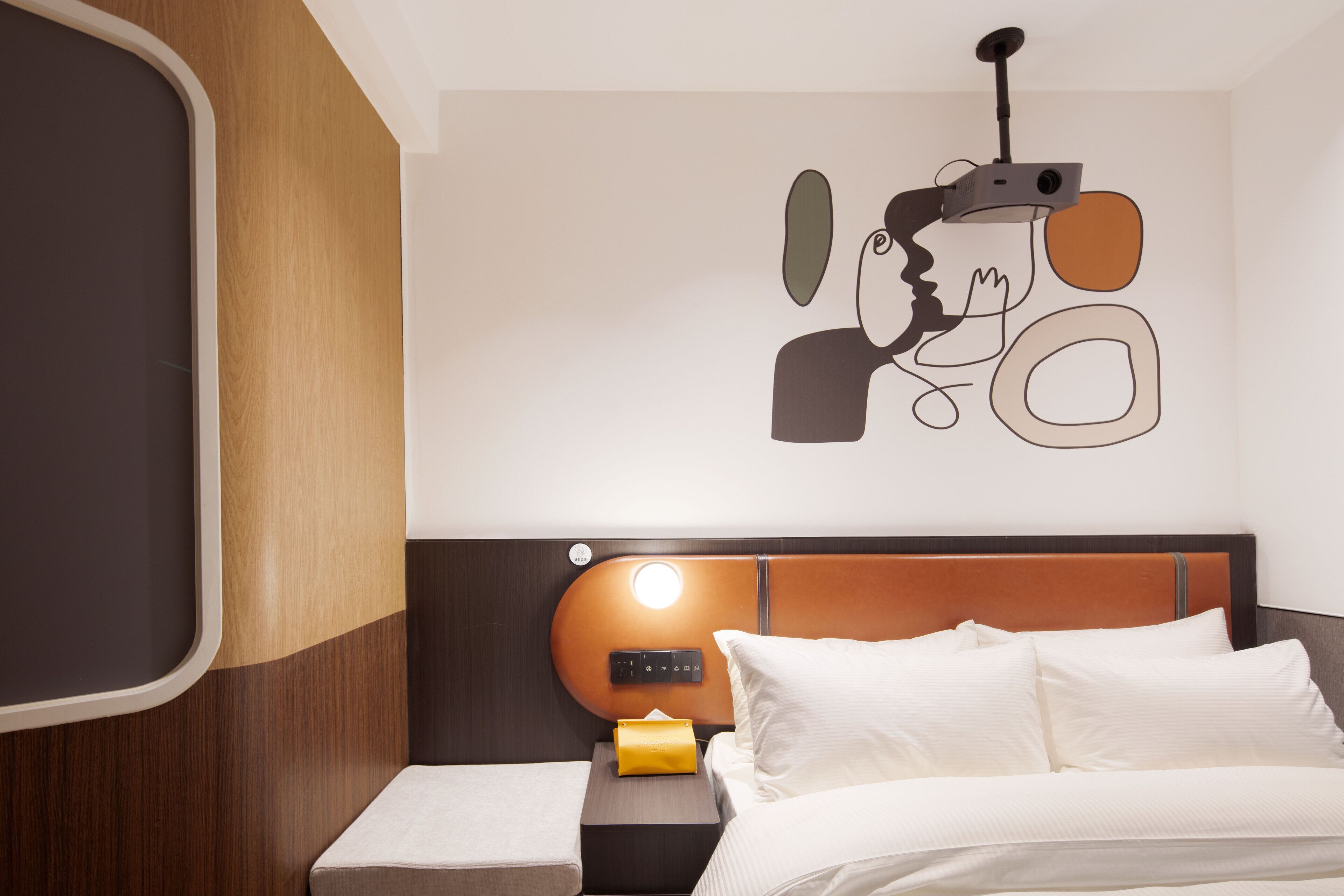 Une chambre d’hôtel moderne avec une tête de lit en bois, un lit avec du linge de maison blanc, une table de chevet avec une lampe et une œuvre d’art accrochée au mur.