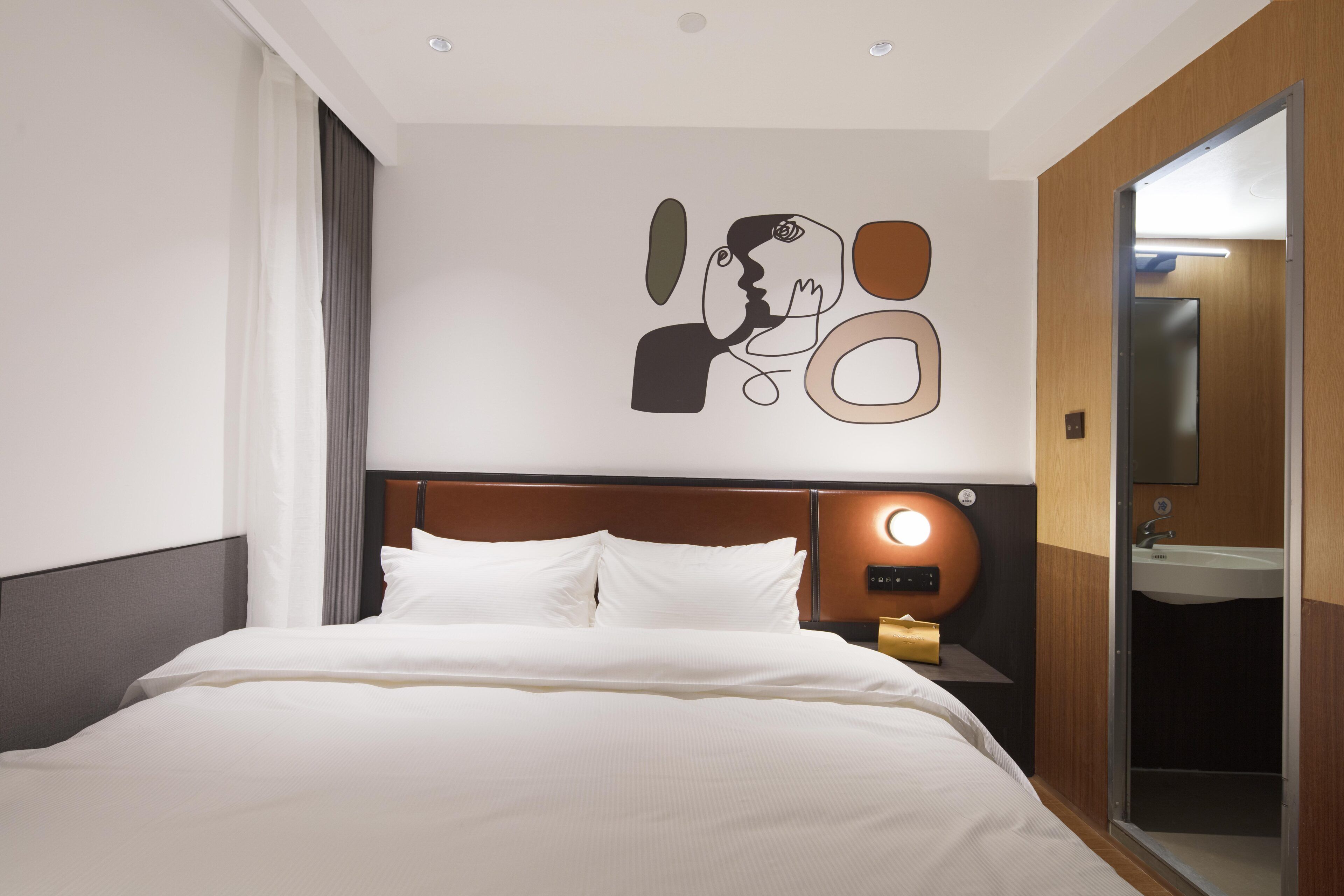 A hotel room with a large bed, a wooden headboard, a wall-mounted artwork, a bedside table with a lamp, and a bathroom with a mirror and sink.