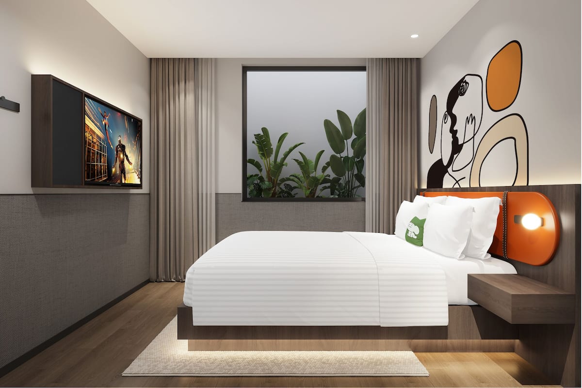 A modern hotel room with a large bed, a window with a view of plants, a wall-mounted TV, and a unique wall art piece.