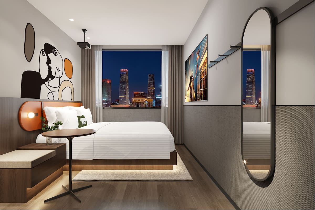A modern hotel room with a large bed, a bedside table, a round mirror, and a city view.