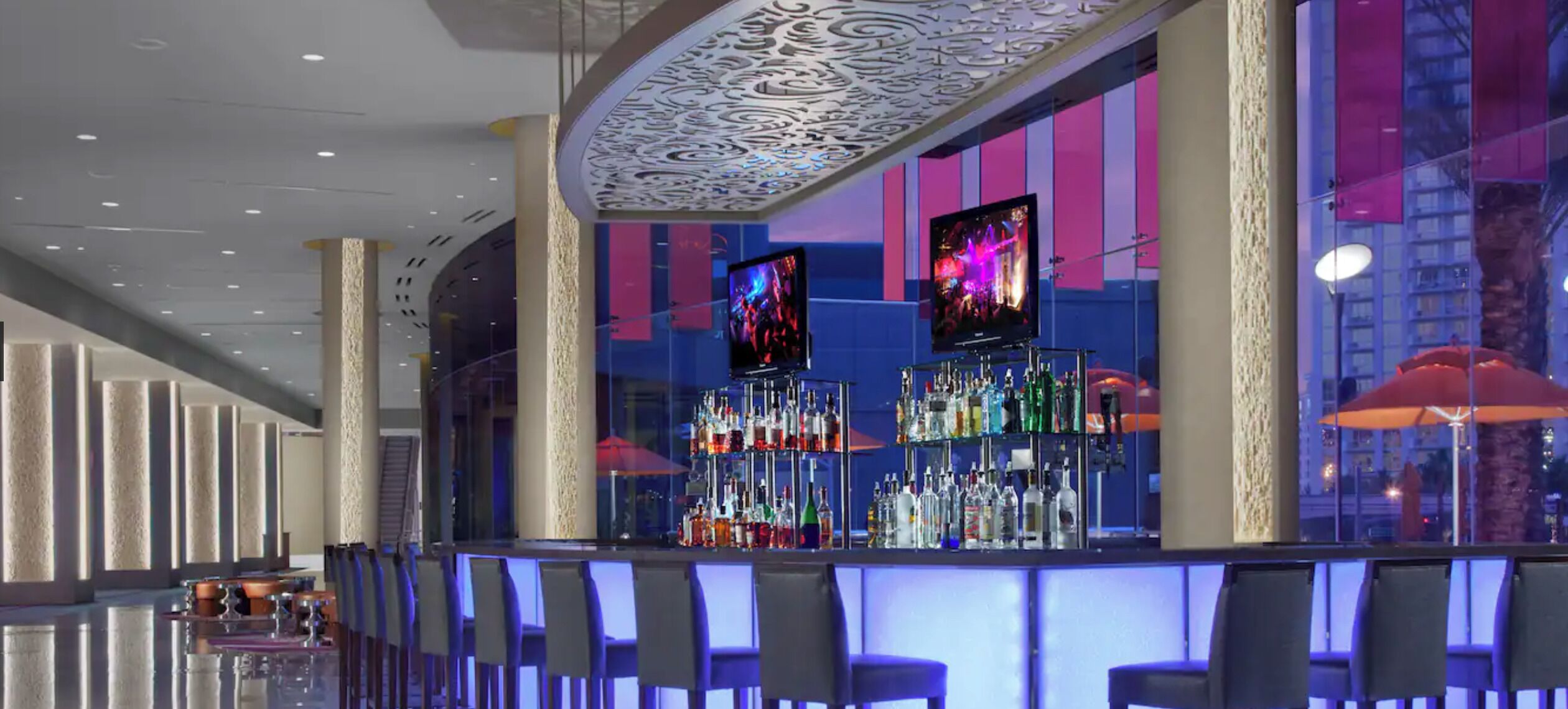 A modern bar area with a glass partition, multiple liquor bottles, and two screens displaying visuals.