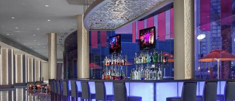 A modern bar area with a glass partition, multiple liquor bottles, and two screens displaying visuals.