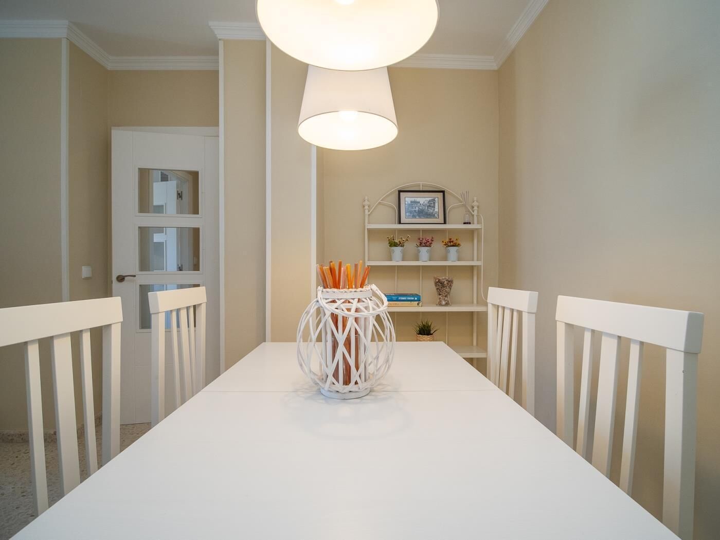 Un comedor con una mesa blanca, sillas, una lámpara colgante y un estante con objetos decorativos.