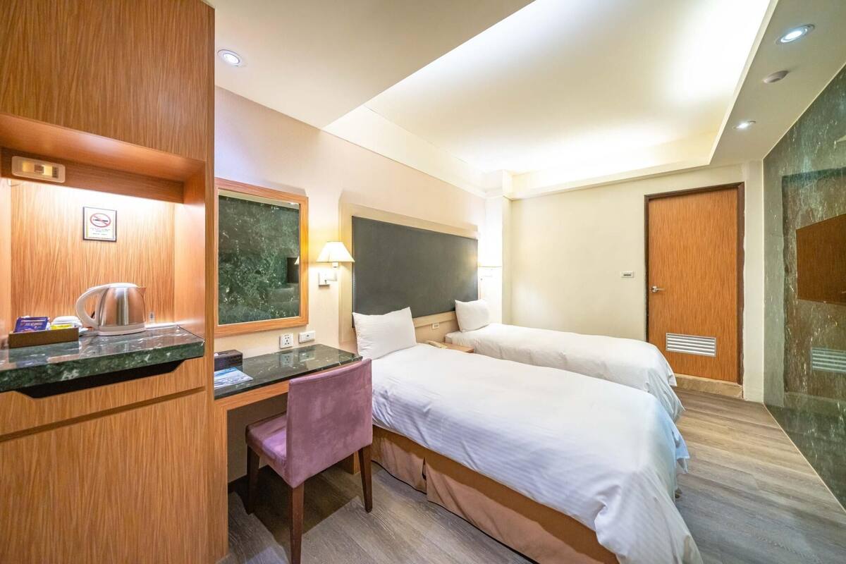 Ein Hotelzimmer mit zwei Betten, einem Schreibtisch mit Stuhl, einem kleinen Tisch mit Wasserkocher, einem Spiegel und einer Tür.