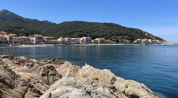 A coastal town with buildings along the shore, rocky foreground, and mountains in the background.
