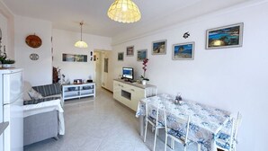 A spacious living area with a dining table, sofa, and TV. There are decorative items and paintings on the walls.