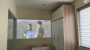 Eine projizierte Leinwand zeigt eine Wiedergabeliste mit Songtiteln und Künstlern in einem Raum mit einem Holzkasten und einem Fenster mit Jalousien.