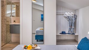 A modern hotel room with a large mirror, a bed with a blue headboard, a small table with a tray of fruit and a drink, and a walk-in closet with hanging clothes.
