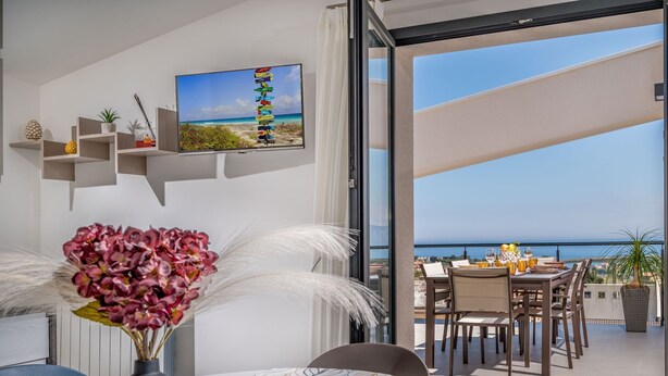 A modern interior with a dining area, a view of the sea, and a TV displaying a beach scene.