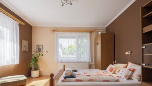 Une chambre avec un lit en bois, une table de chevet, un radiateur, une fenêtre avec des rideaux, une plante et une étagère fixée au mur.