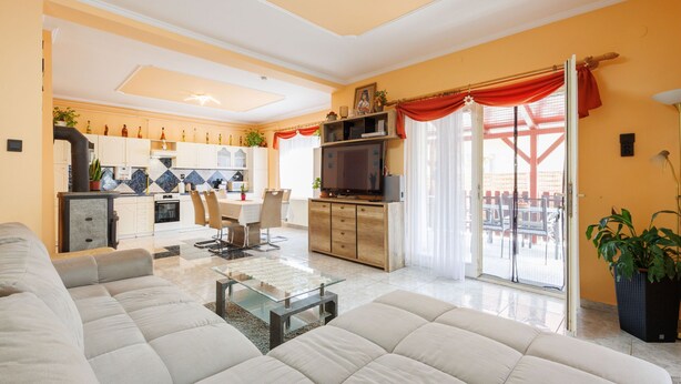 Un salon moderne avec un grand canapé beige, une table basse en verre et un téléviseur. On aperçoit la cuisine au fond, avec des meubles blancs et un rideau rouge.