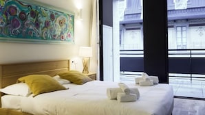 Et hotelværelse med en stor seng, et billede på væggen og udsigt til en bygning gennem vinduet.