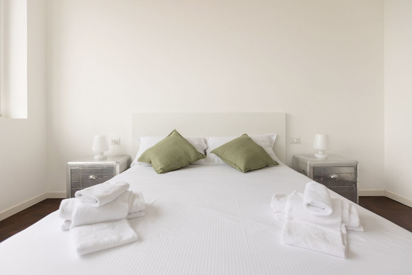 Cama bem arrumada com lençóis brancos e duas almofadas verdes, com duas mesas de cabeceira com luminárias brancas e toalhas brancas dobradas.