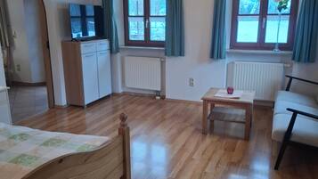 Ein Zimmer mit Holzboden, einem Bett, einem Schreibtisch, einem Stuhl und zwei mit Vorhängen versehenen Fenstern.
