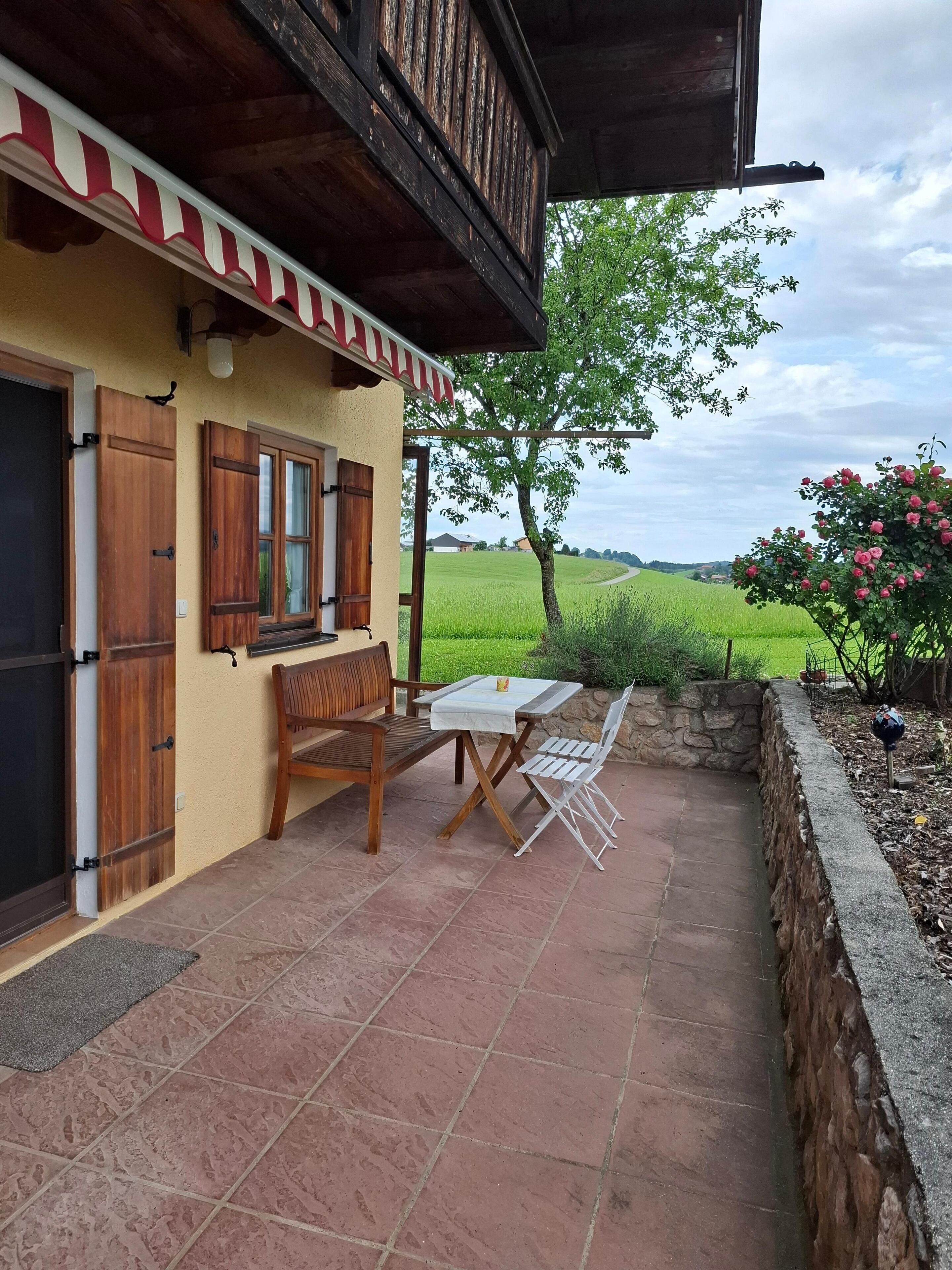 An outdoor patio with a wooden table and chairs, a stone wall, and a view of a green field.
