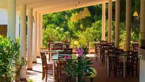 An outdoor patio area with tables and chairs, surrounded by greenery and columns.