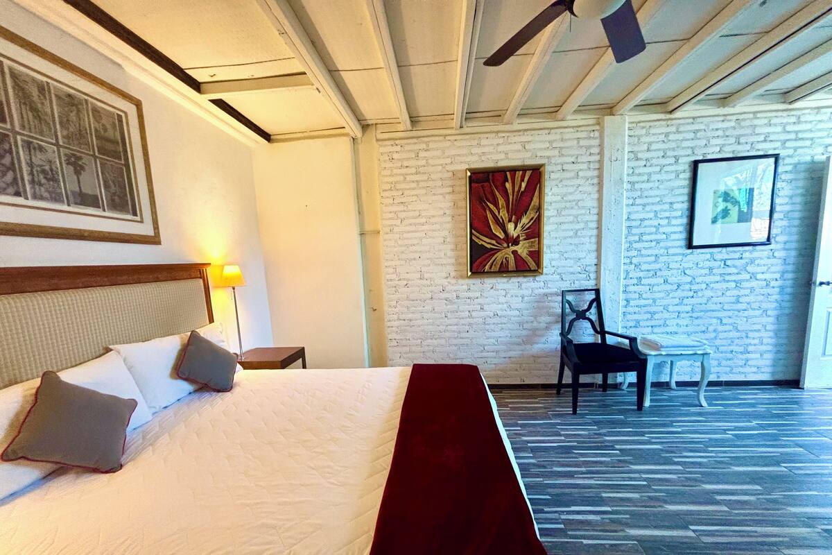 A hotel room with a large bed, a wooden headboard, a red throw blanket, a nightstand with a lamp, a chair, a small table, and a brick wall with artwork.