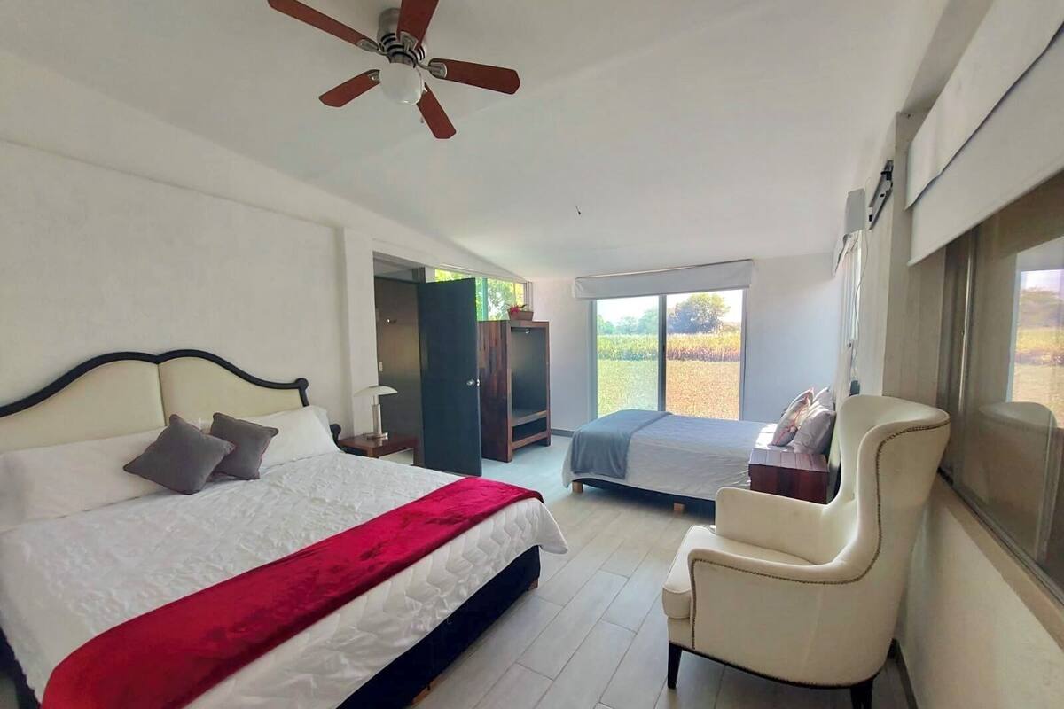 A bedroom with a bed, a chair, a ceiling fan, and a large window.