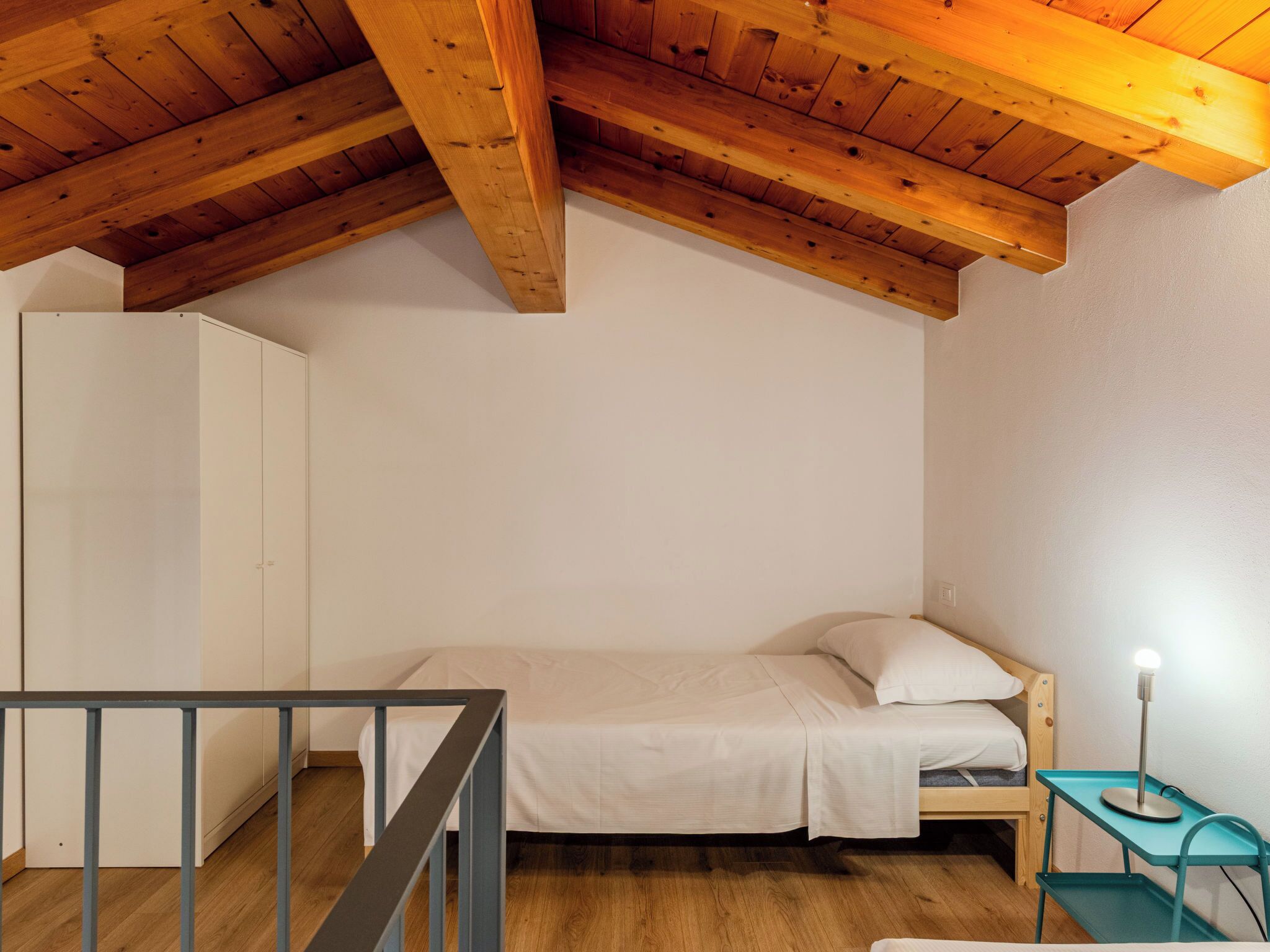 A small, single-bed room with a sloped wooden ceiling, a white wardrobe, and a small table with a lamp.