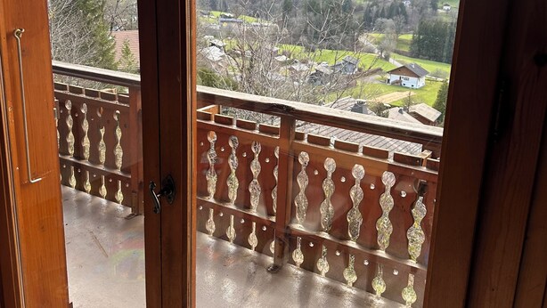 En träbalkong med utsikt över ett bergigt landskap och ett litet hus i fjärran.