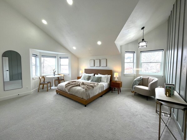 A bedroom with a large bed, two bedside tables, a chair, and a desk. There are windows on two walls, and a chandelier hangs from the ceiling.