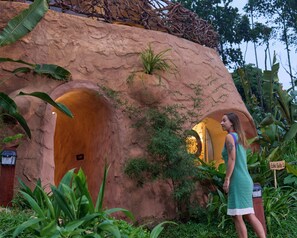 A person standing in front of a rustic, cave-like structure with arched doorways, surrounded by lush greenery. - Toroland Munnar  (Devikolam)