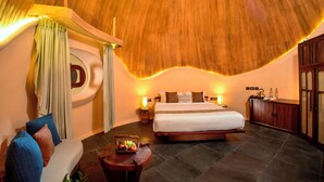 A bedroom with a large bed, a wooden ceiling, a round mirror, a bedside table with a lamp, and a small table with fruits.
