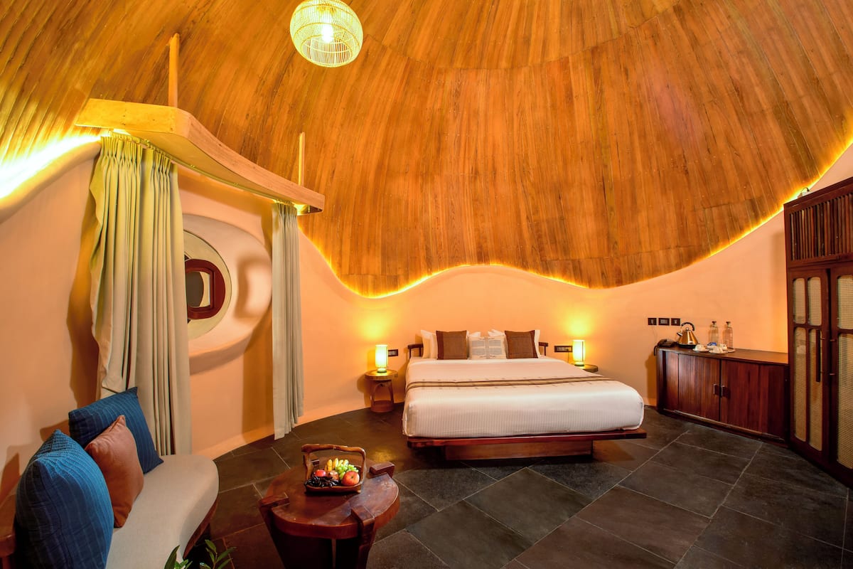 A bedroom with a large bed, a wooden ceiling, a round mirror, a bedside table with a lamp, and a small table with fruits.
