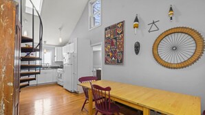 Una cocina con armarios blancos, una mesa de madera y una silla roja. Hay un ventanal grande sobre el fregadero y un cuadro abstracto colorido en la pared.