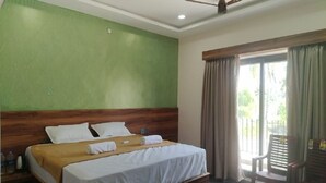A hotel room with a bed, a wooden headboard, a ceiling fan, a window with curtains, and a chair by the window. - Trigon Retreat (Puducherry)