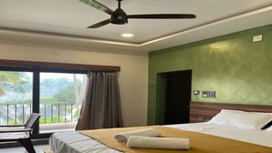 A hotel room with a bed, a chair, a ceiling fan, and a view of the outdoors. - Trigon Retreat (Puducherry)