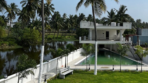 A modern two-story building with a pool, surrounded by palm trees and a body of water. - Trigon Retreat (Puducherry)