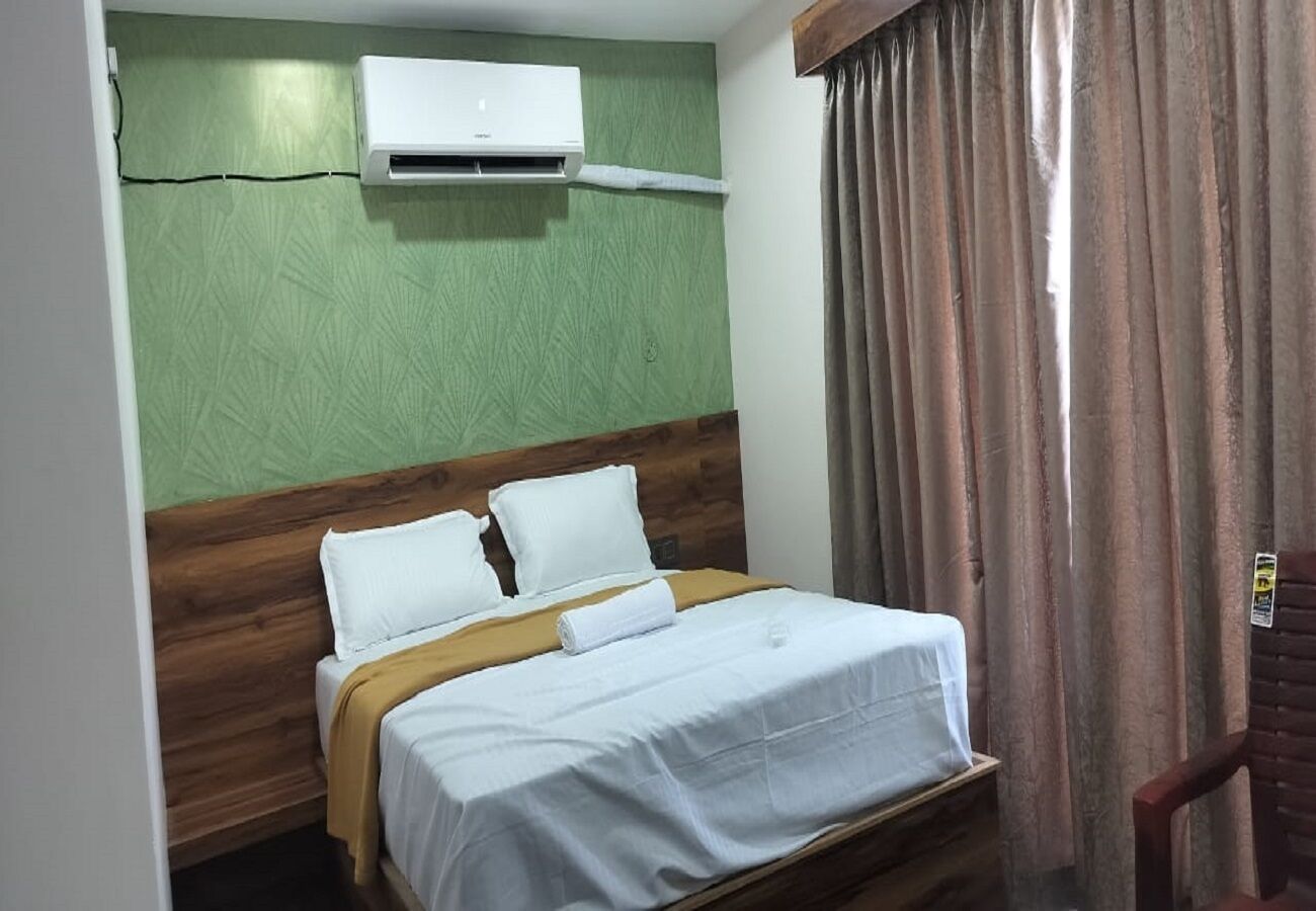 A hotel room with a bed, a wooden headboard, a green textured wall, an air conditioning unit, and a curtain.