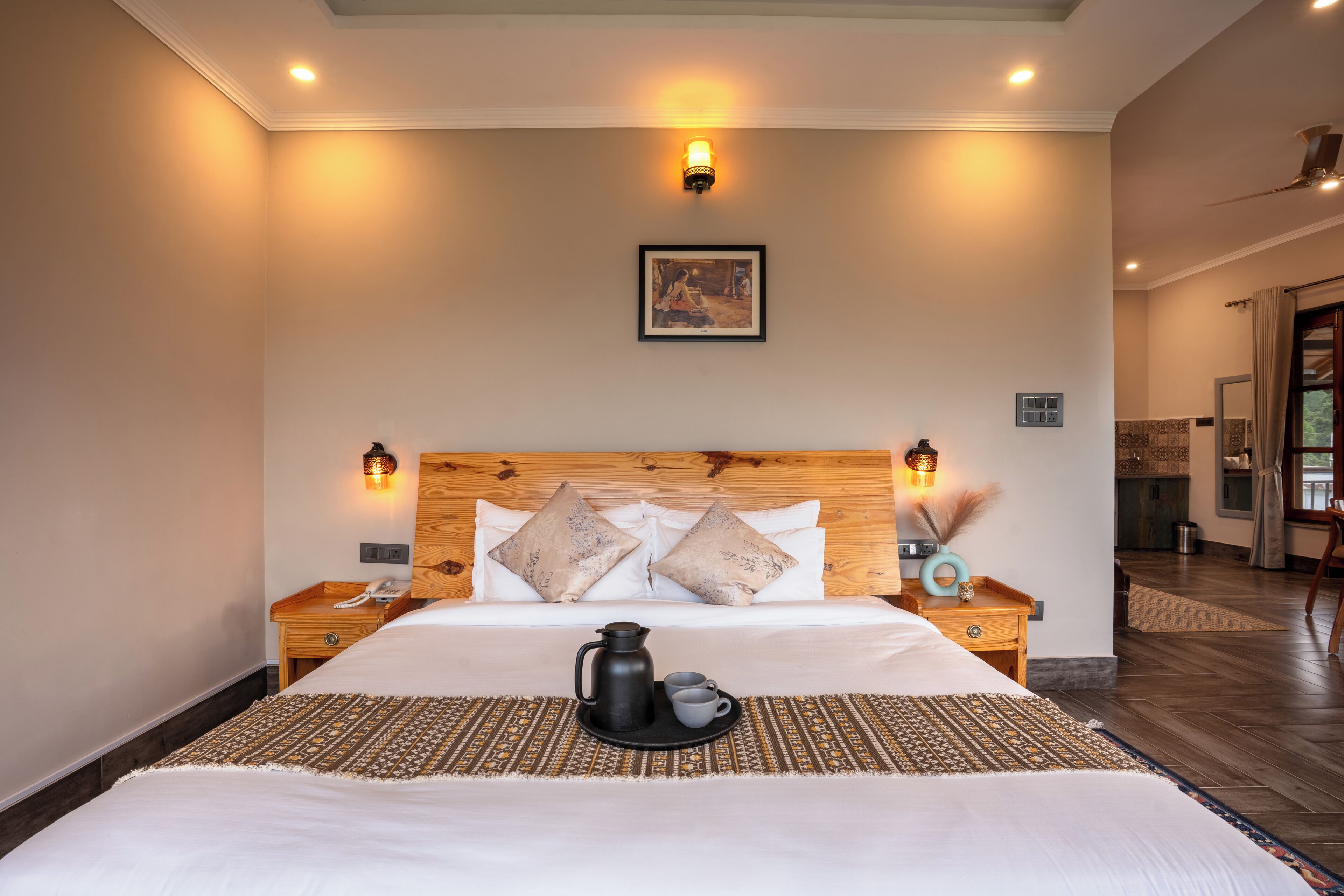 Habitación de hotel con una cama grande, cabecera de madera, lámparas de noche, un cuadro en la pared y una bandeja con cafetera y taza.