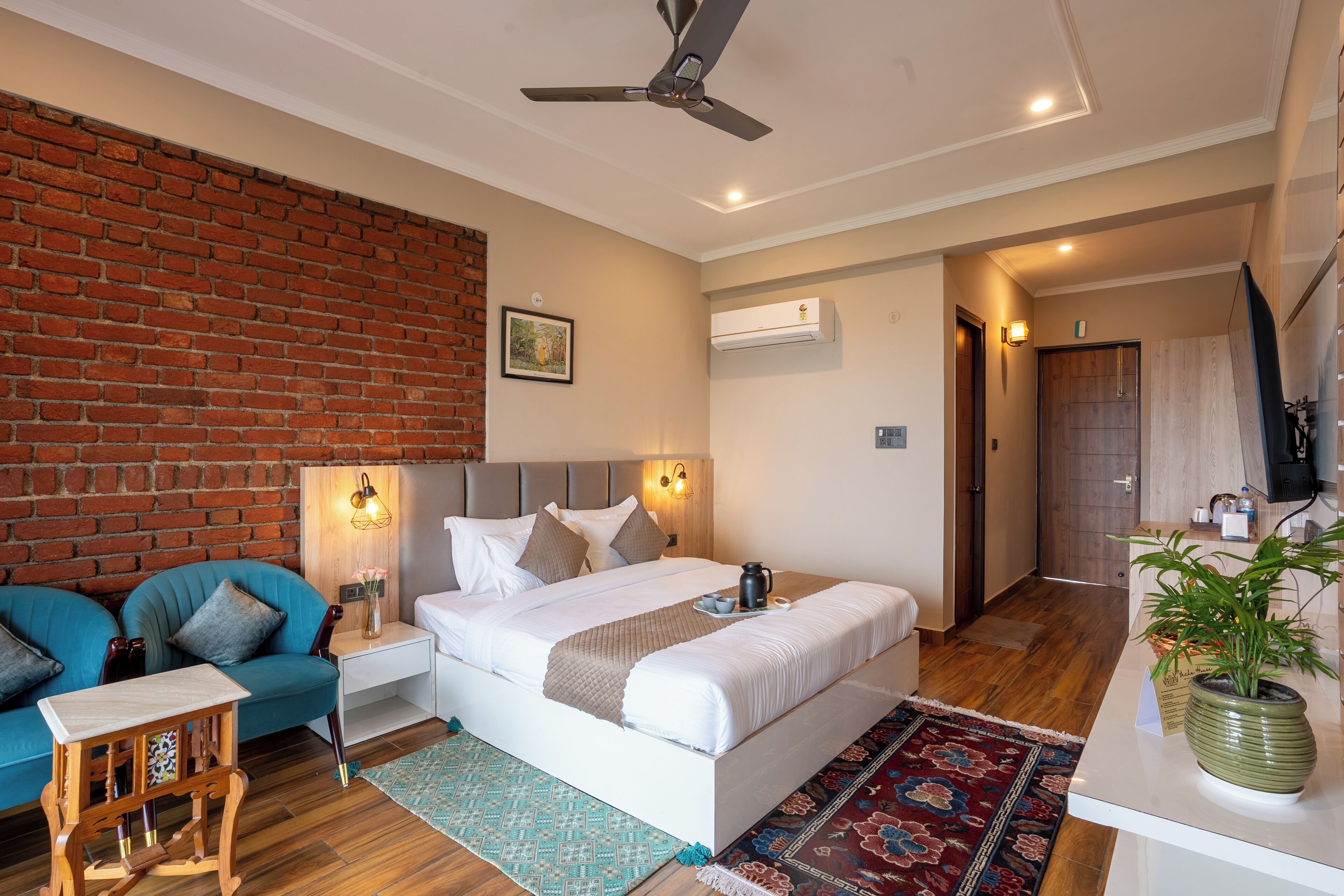 A modern bedroom with a brick wall, a bed with white linens, a blue sofa, a small wooden table, and a potted plant.