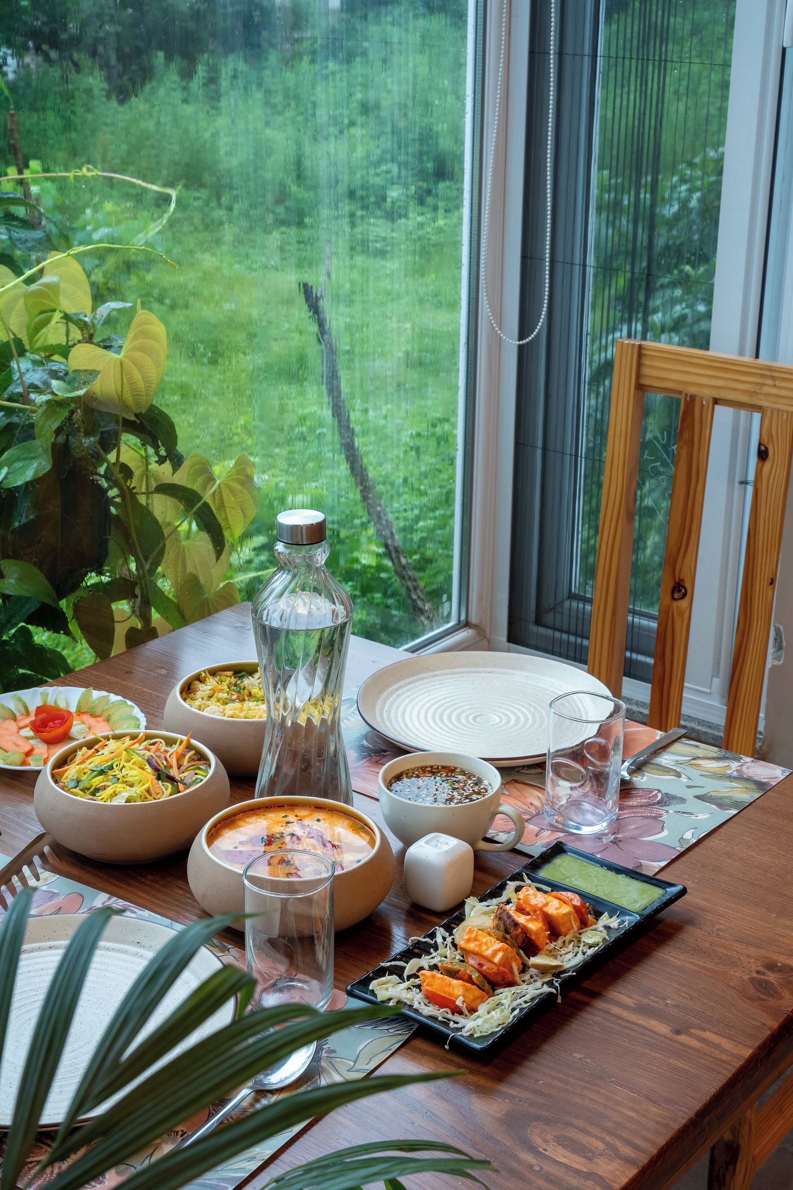 A dining table with various dishes, a glass bottle, and a view of greenery through a window.