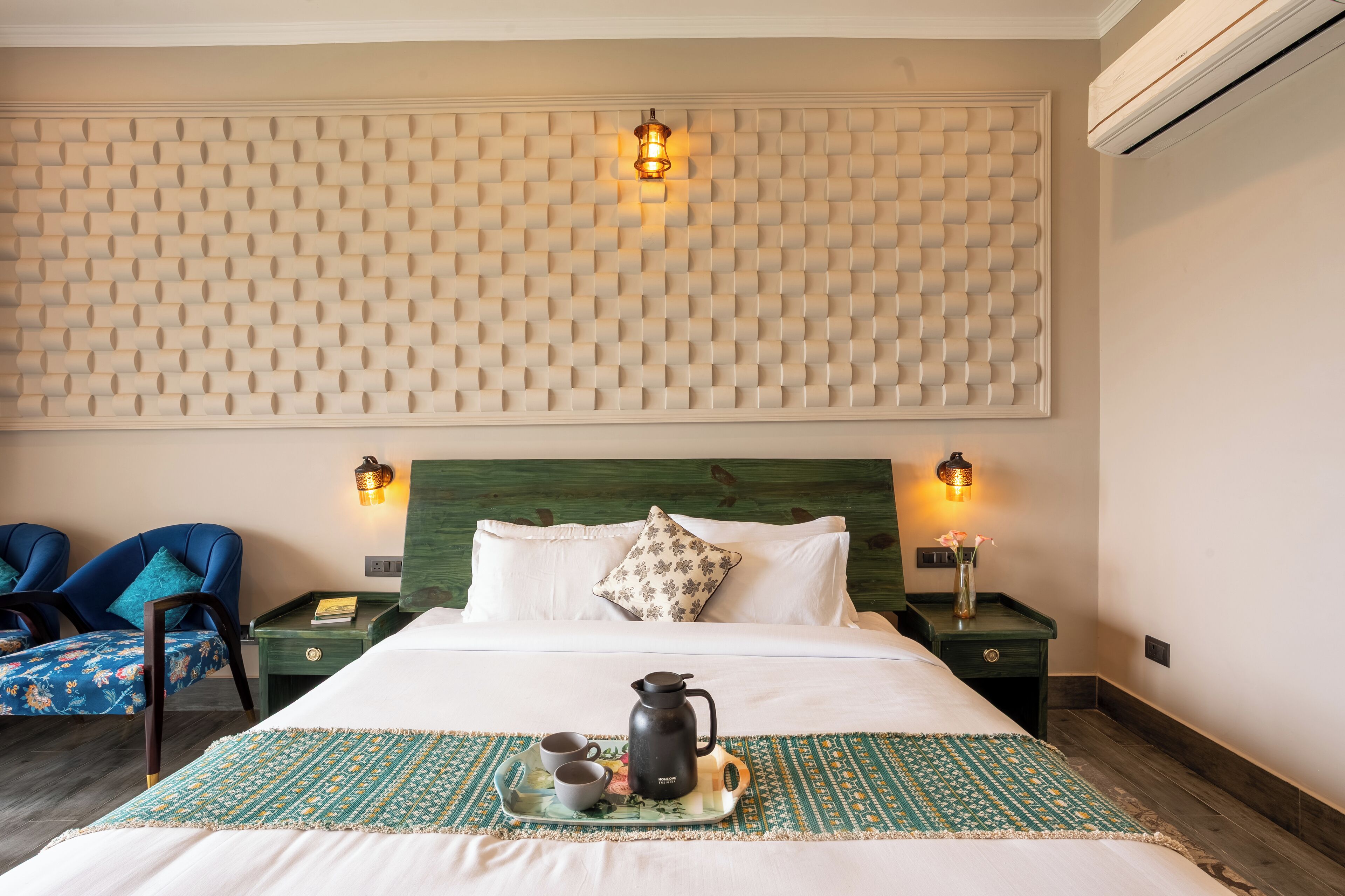 A hotel room with a bed, two chairs, a nightstand with a lamp, and a tray with a coffee pot and cups.