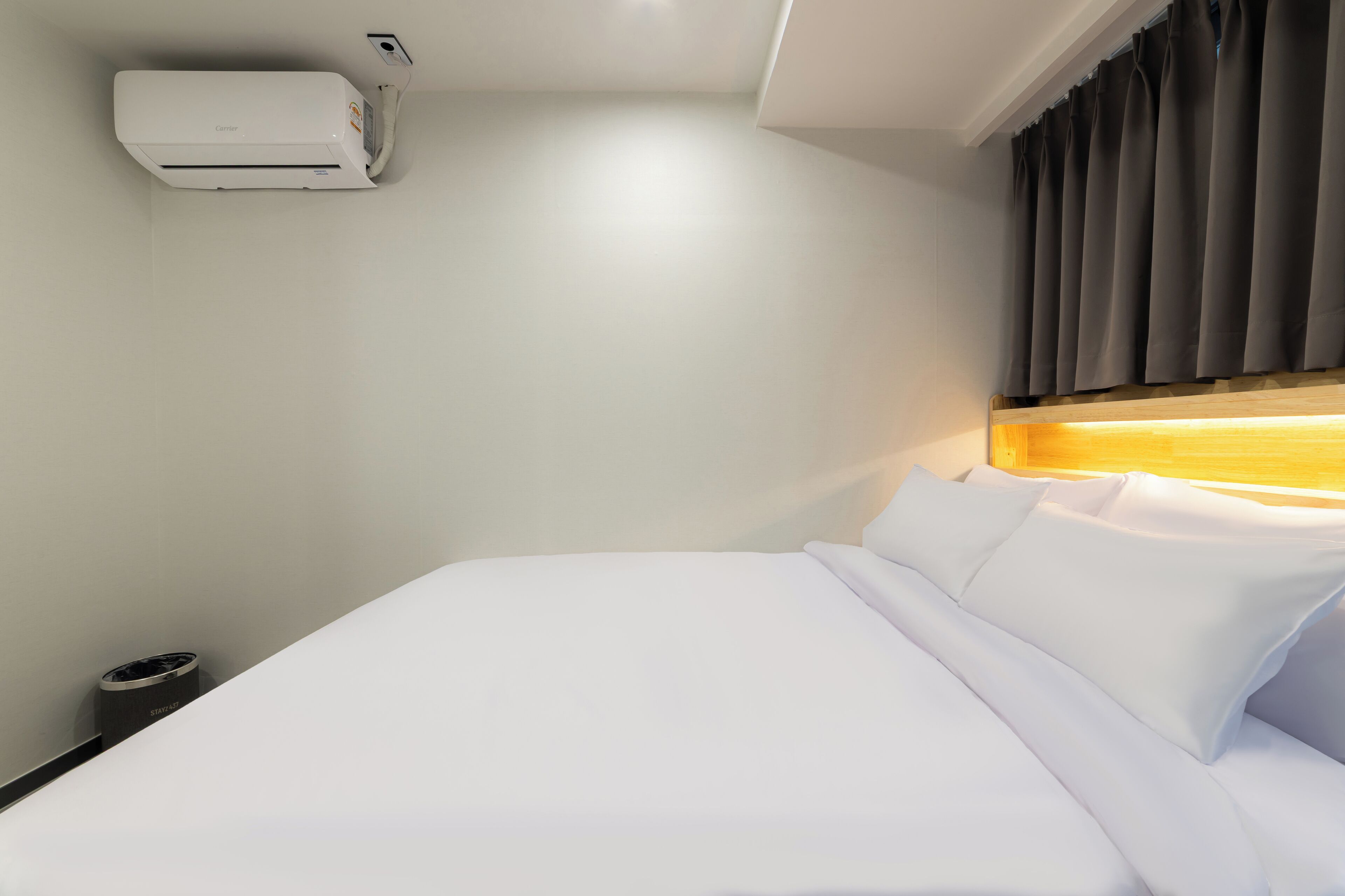 A hotel room with a bed, a wall-mounted air conditioner, a remote control, and a window with curtains.