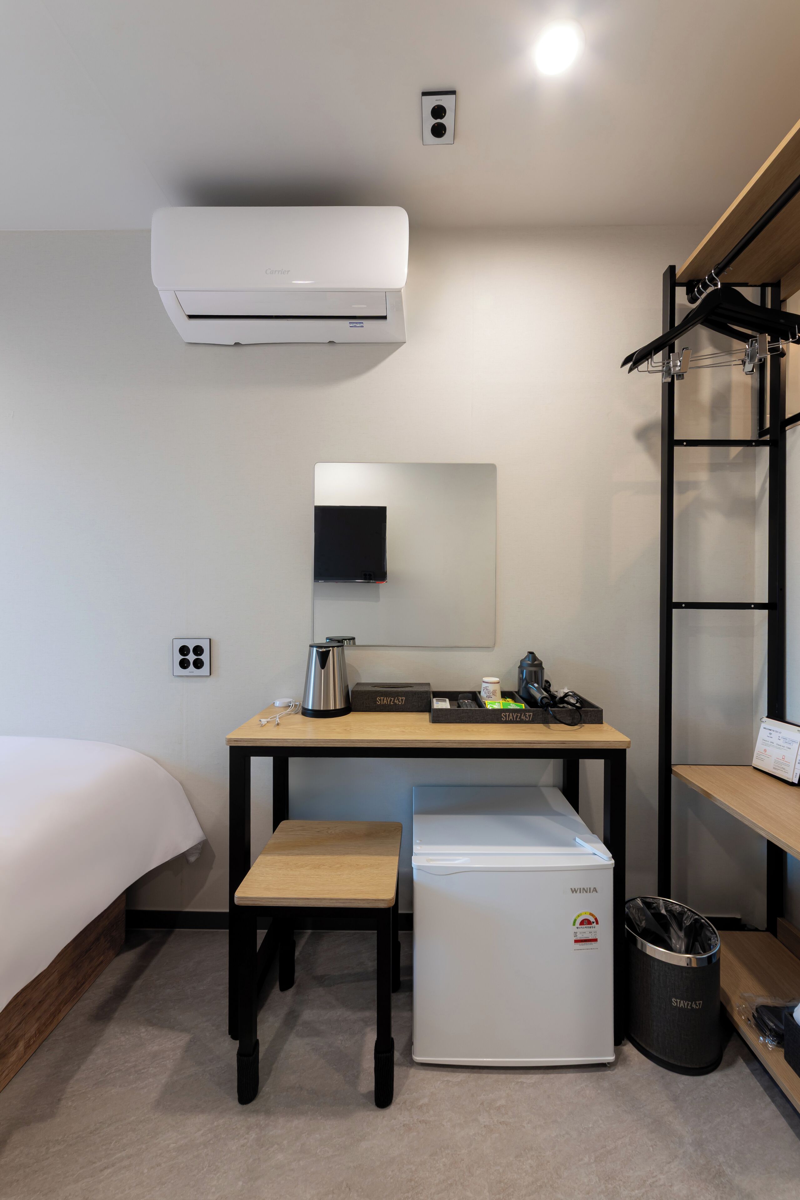 A compact hotel room with a bed, a desk with a computer, a small fridge, and a wall-mounted air conditioner.