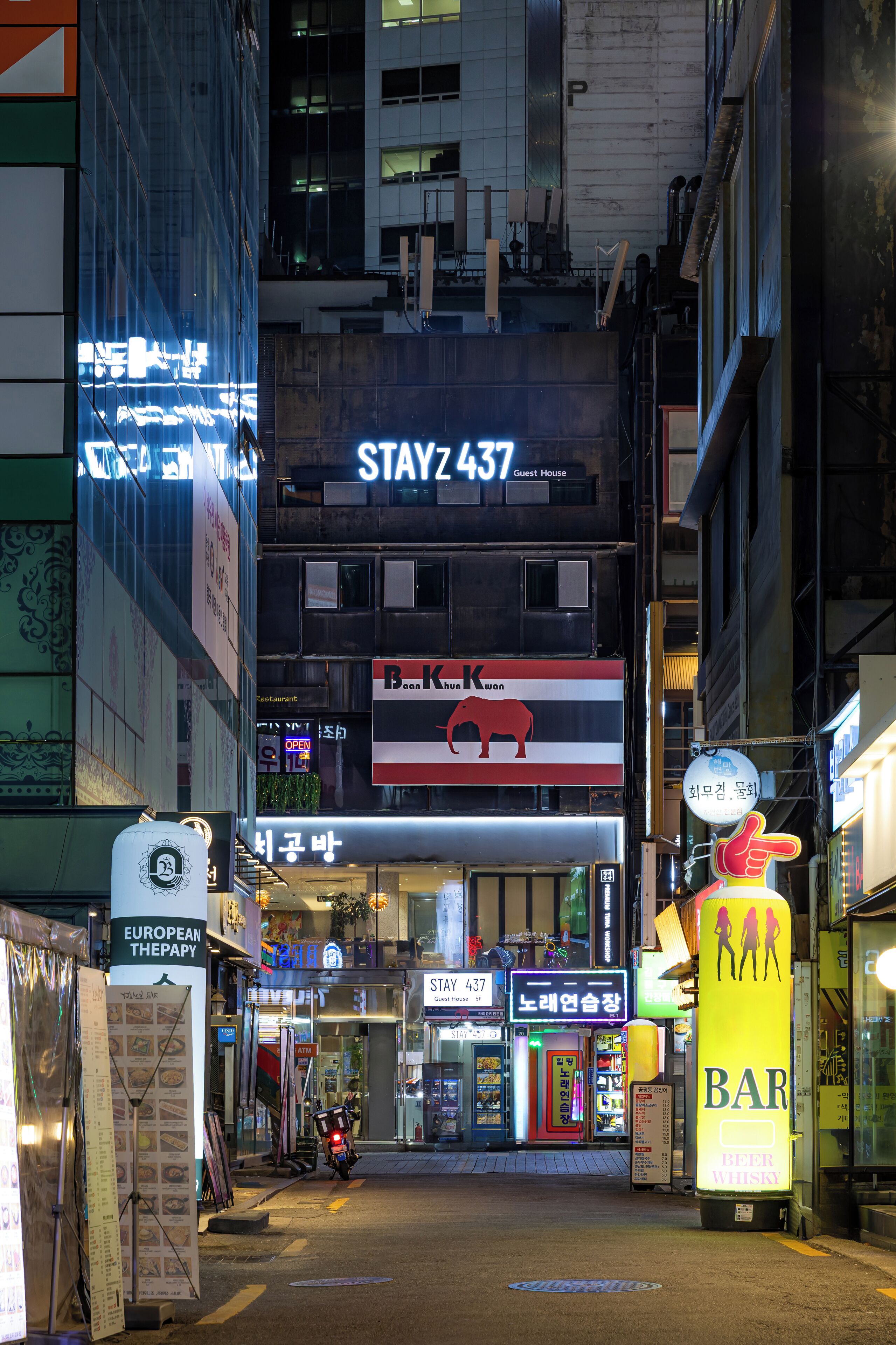Una calle urbana iluminada con un letrero destacado que dice 'STAYZ 437', varios comercios y un gran letrero amarillo que dice 'BAR'.