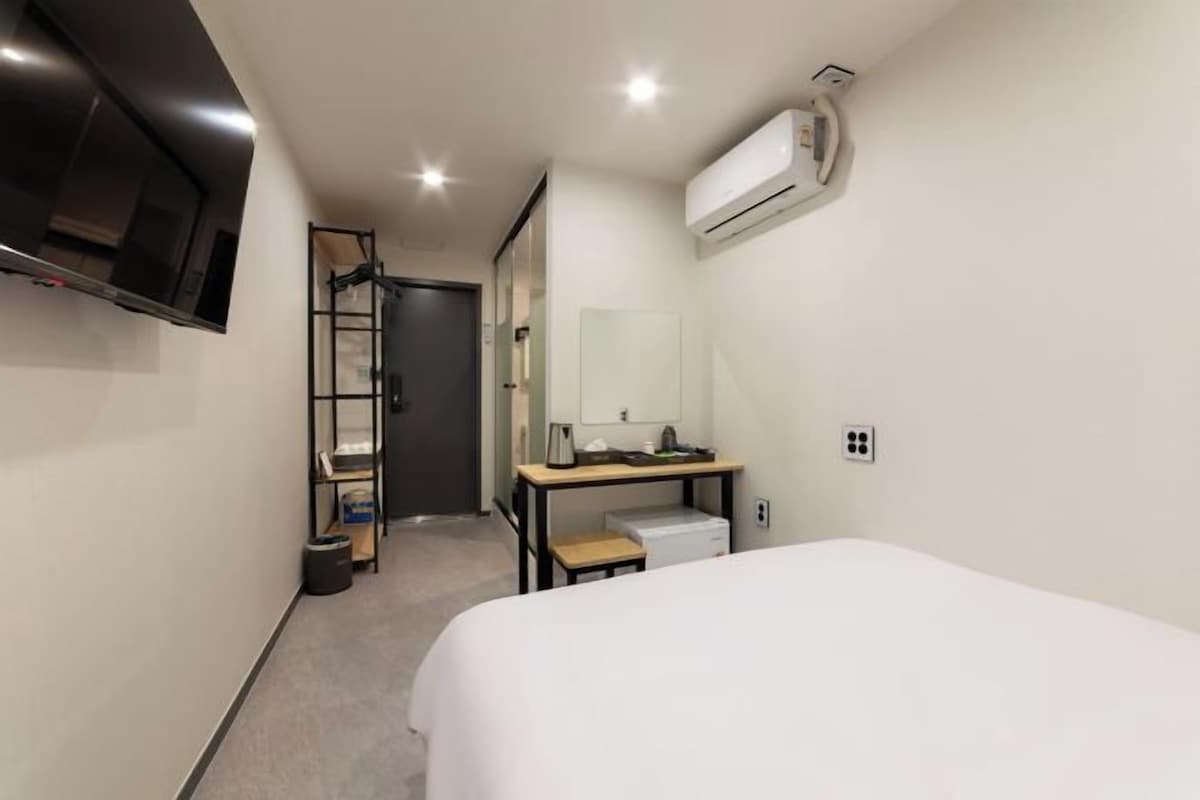 A hotel room with a bed, a desk with a mirror, a TV mounted on the wall, and an air conditioning unit.