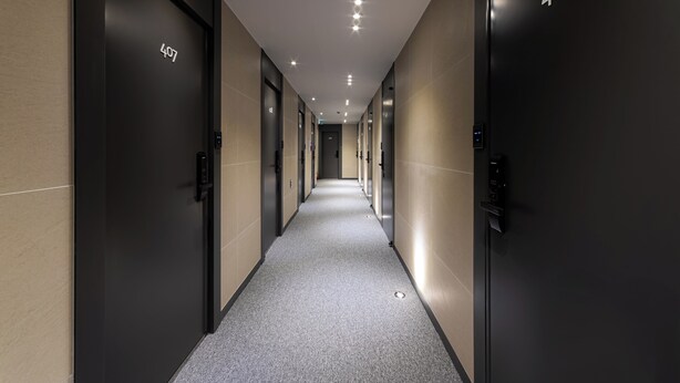 En modern hotellkorridor med numrerade dörrar, ett heltäckningsmatta och infälld belysning.
