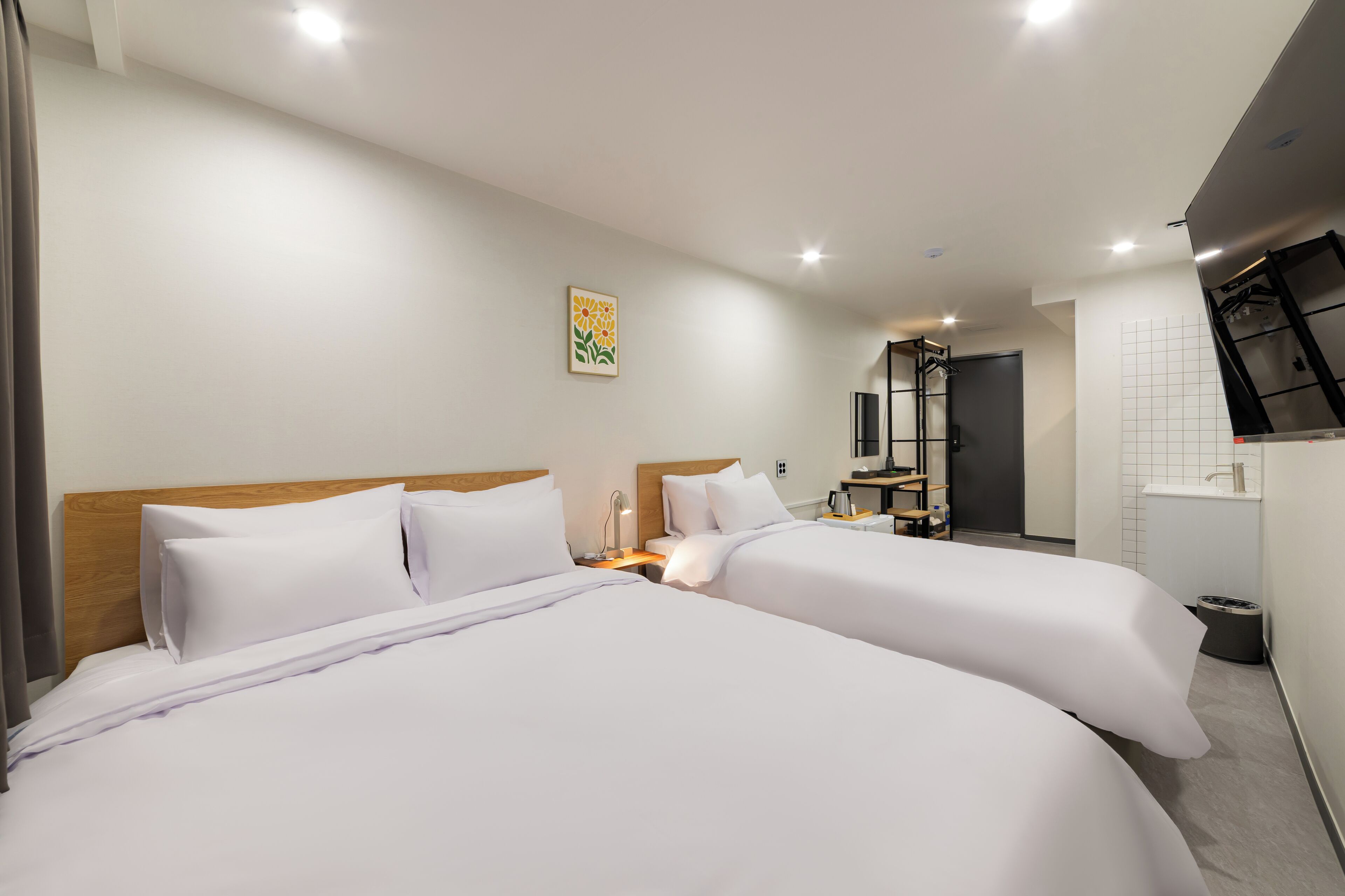 Habitación de hotel con dos camas, cabecero de madera, mesita de noche con lámpara, pequeño escritorio con silla, espejo y vista al baño.