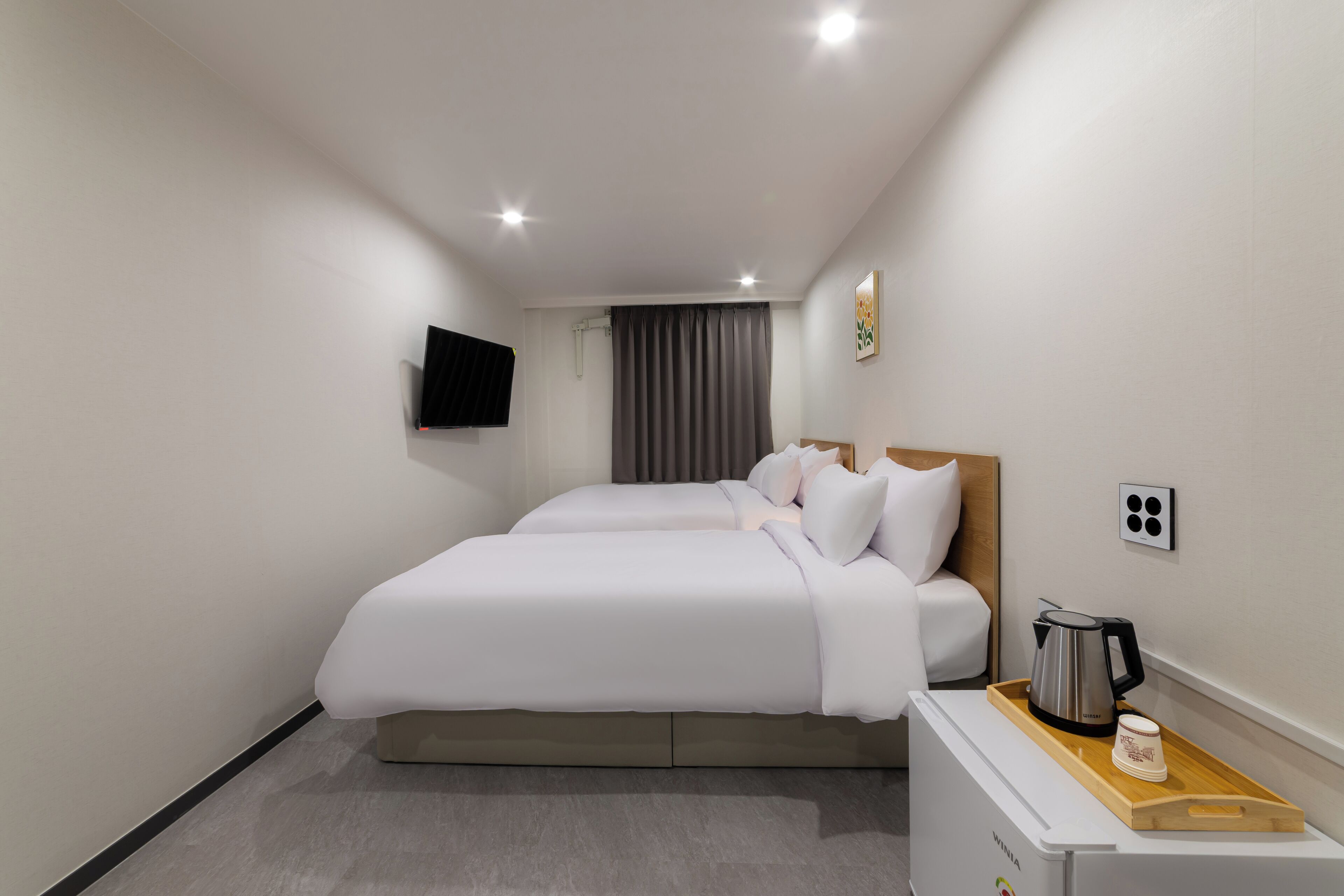 Quarto de hotel moderno com uma cama grande, mesa de cabeceira com chaleira e xícaras, e televisão de tela plana na parede.