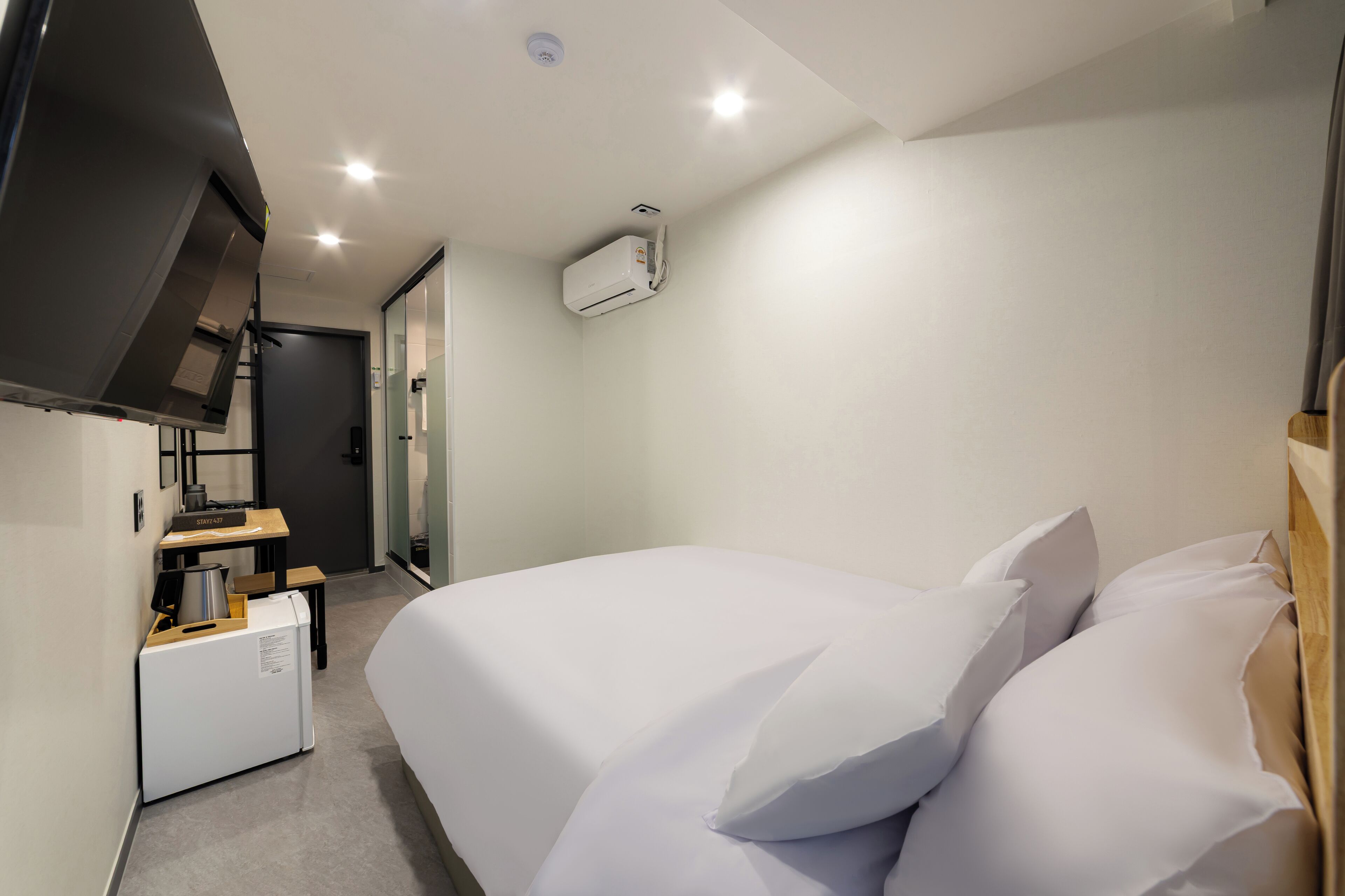 A hotel room with a bed, a small kitchenette, and a ceiling-mounted air conditioner.