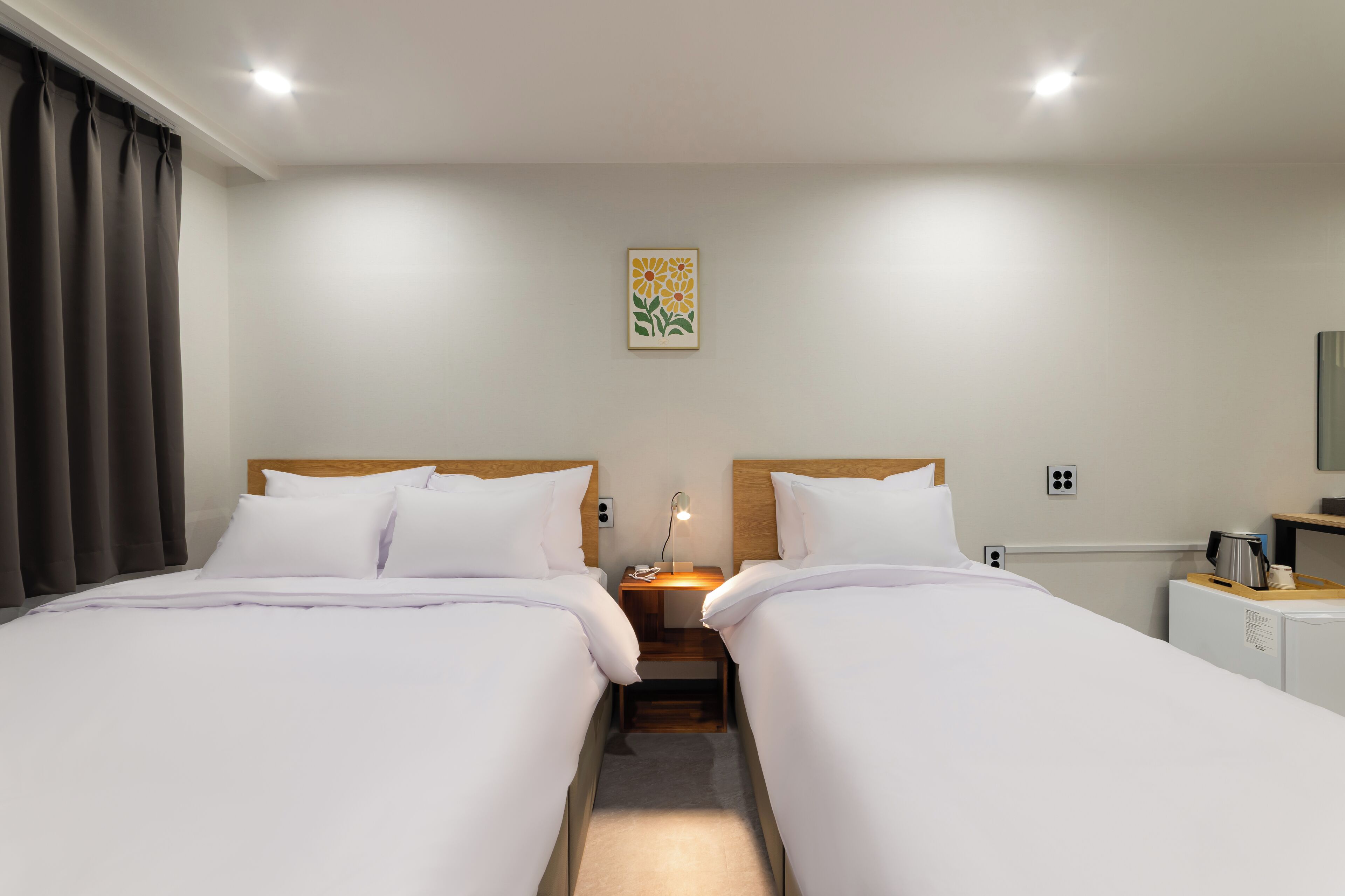 Une chambre d’hôtel avec deux lits, une table de chevet avec une lampe, une petite table avec une bouilloire et un tableau au mur.
