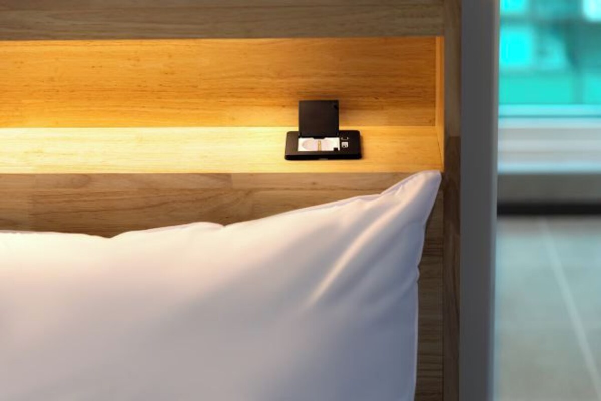 A bed with a white pillow and a black object on the headboard.