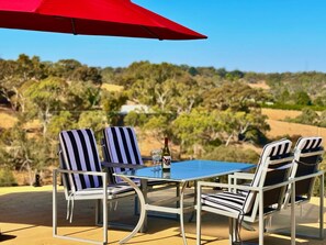 Une table entourée de quatre chaises à rayures et d’un parasol rouge, installée à l’extérieur, avec vue sur les arbres et les collines.