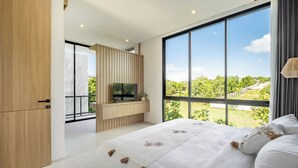 Ein modernes Schlafzimmer mit einem großen Bett, einem an einer Holzvertäfelung montierten Fernseher und einem Balkon mit Blick auf Grünflächen.